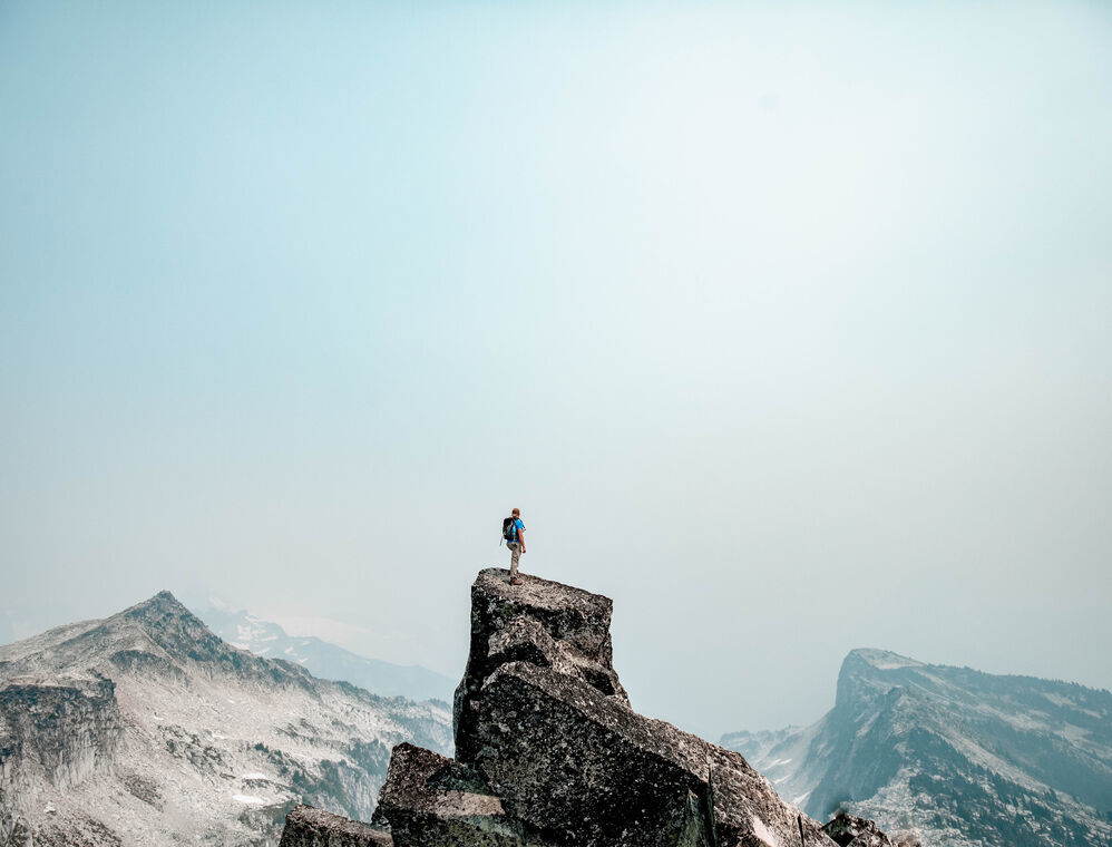 A Person standing on top of a rocky mountain looking into the distance. There are also some other rocky mountains and snow besides.