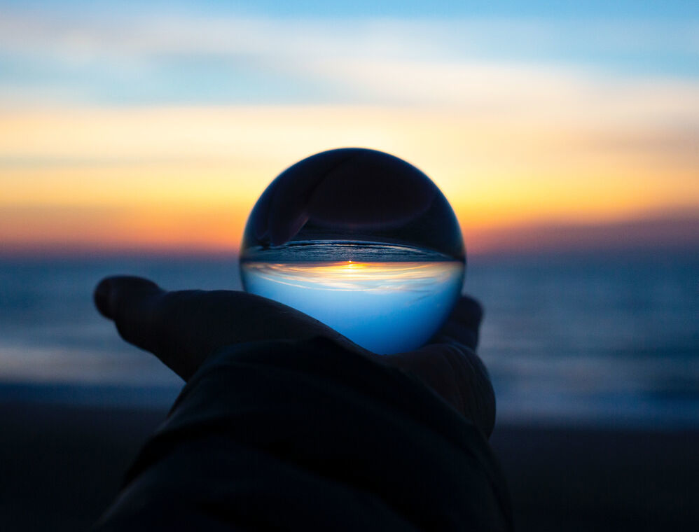 A Hand at arms length, holding a glass globe against the horizon. You can see the sunset above the sea through the globe.