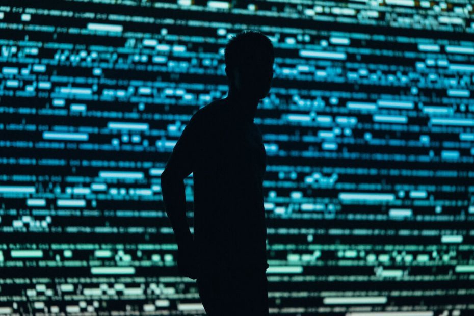 A Person standing in front of a giant screen with unrecognizable data strings in different colour. The person is unrecognizable as well.